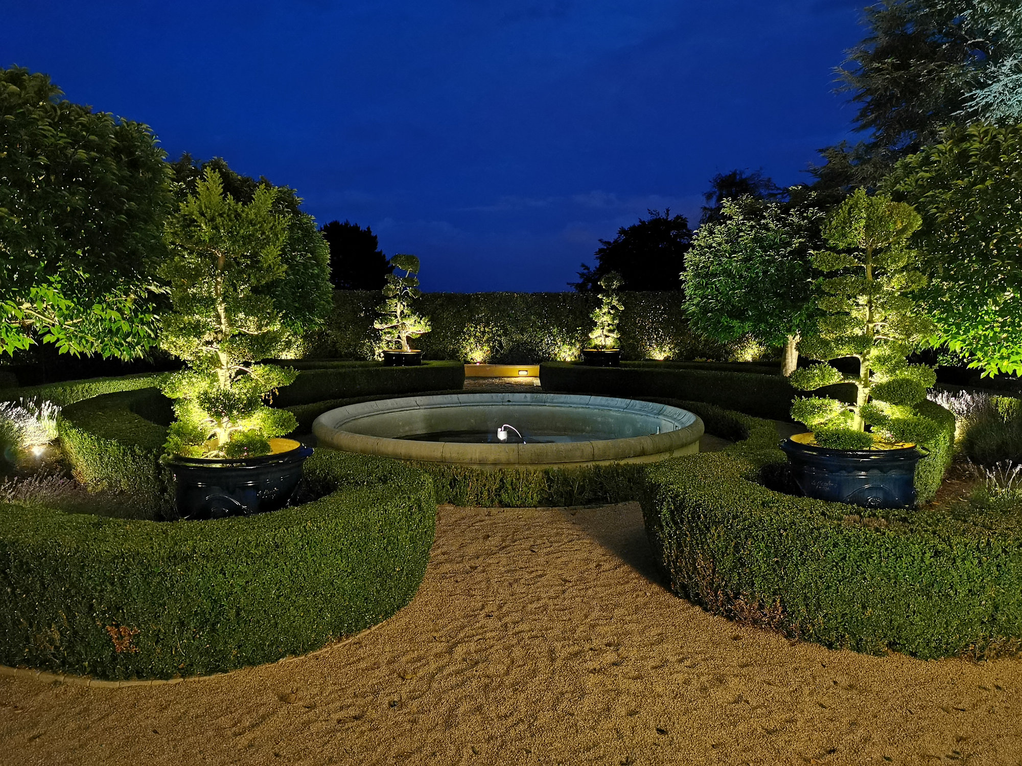 Exklusive Gartenbeleuchtung bei Nacht – beleuchtete Formgehölze rund um einen Brunnen