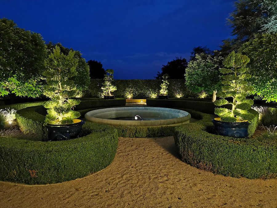 Formgehölze rund um einen Brunnen, stimmungsvoll beleuchtet in der blauen Stunde