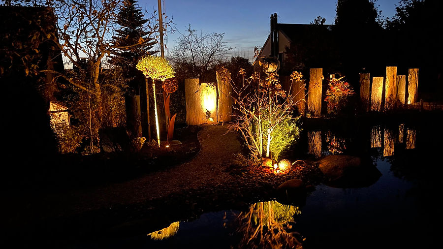 Naturgarten mit Schwimmteich, warm angestrahlten Steinstelen und Gehölzen in der Dämmerung