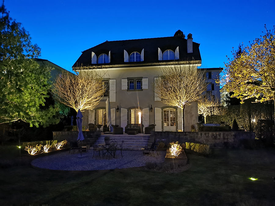 Villa mit angestrahlten Formbäumen, Gartenterrasse und Beetleuchten zur blauen Stunde